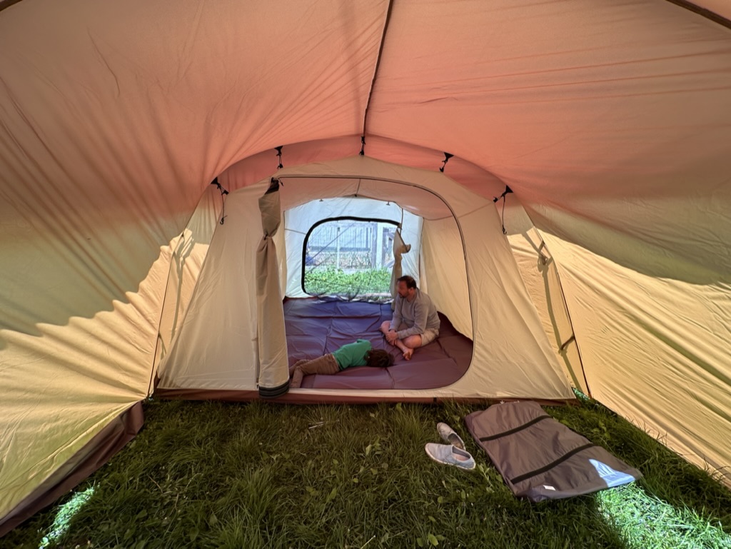 Interior shot of the large tent, taken with a wide angle lens. A smaller room visible in the center/back with a kid and parent in lying on the floor. you can see right through the back screens to the grass beyond.