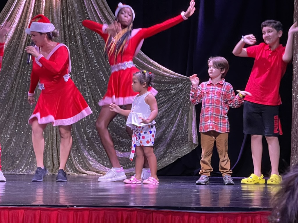 Photo of a group of people on a stage. Two adult women dressed as elves and a bunch of kids aged 5-10 around. Taken mid dance number