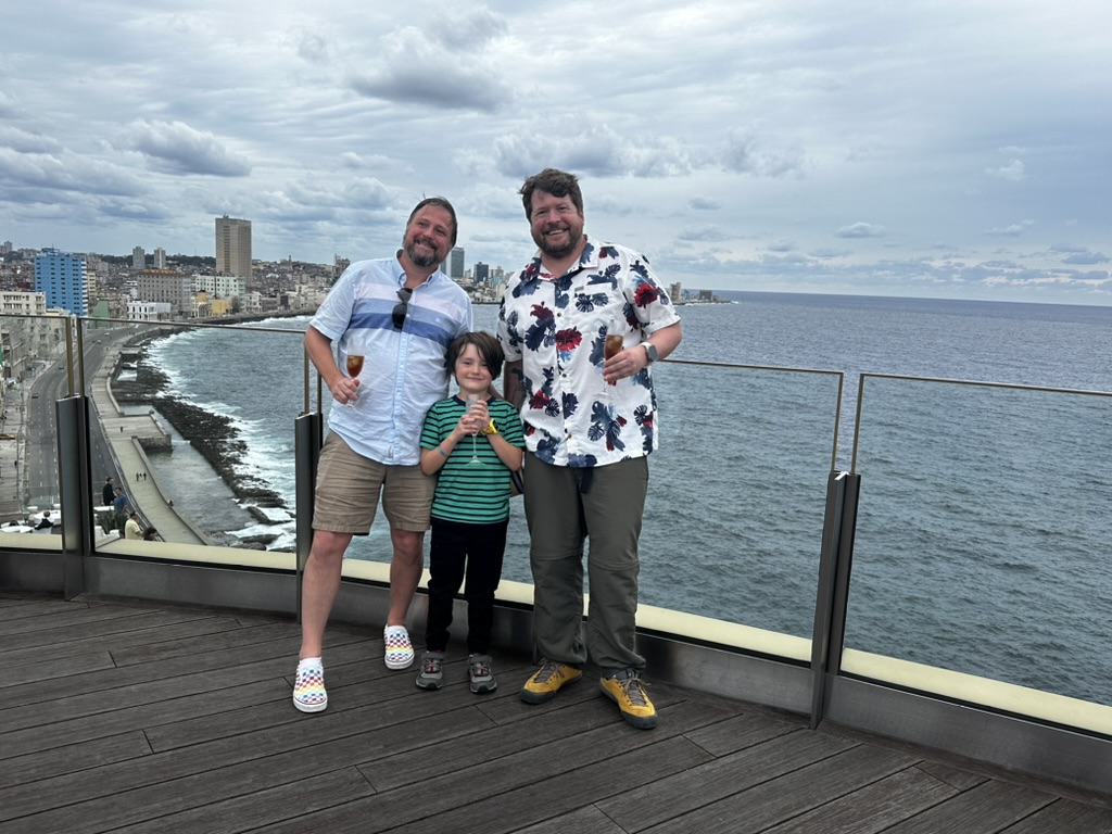 two adults in relaxed summer wear and a kid in a green striped t-shirt lean against a glass railing with the ocean and city of Havana in the backgrounf