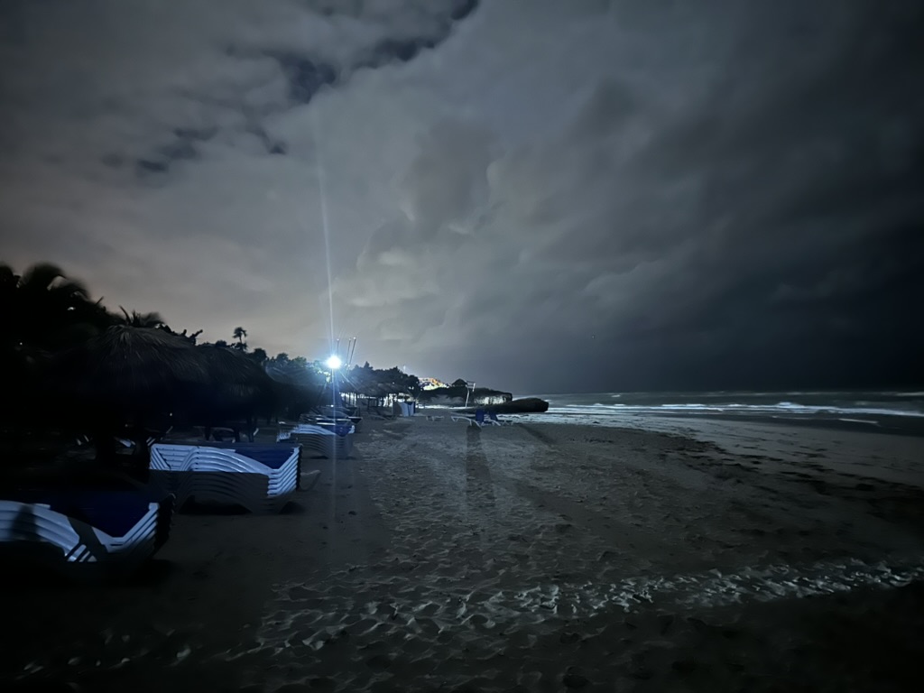 late night photo of the beach. a bight light in the distance and the surf visible.