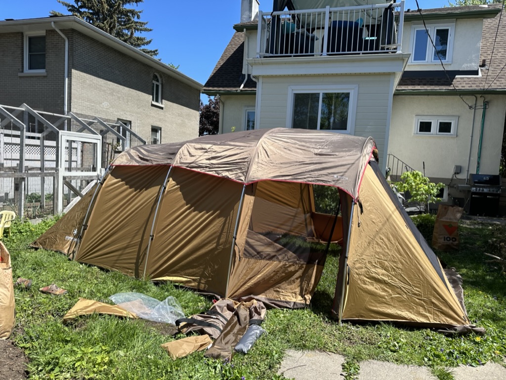 A large brown tent with dark roof is setup in a house's back yard. Green grass, a garden, and several houses are visible