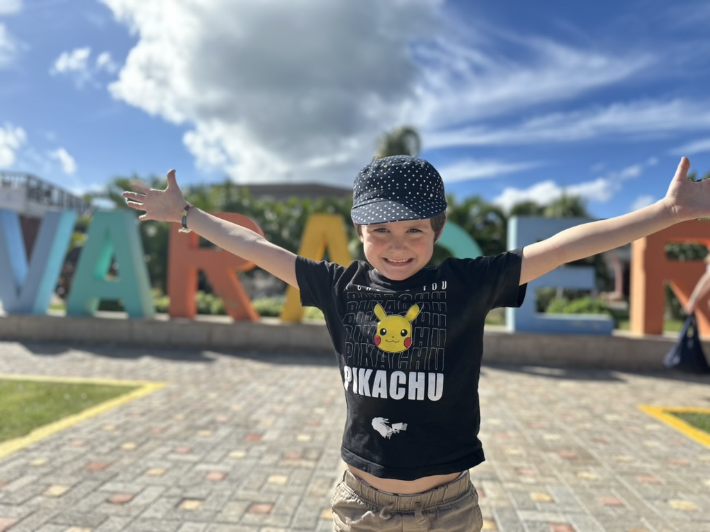 Talia in a black shirt with Pikachu on it with her arms spread wide. In the background a large and multicoloured sign reads Varadero