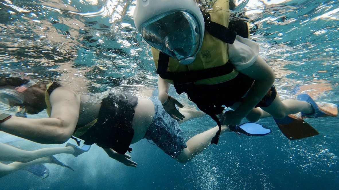 underwater photo taken from below looking up at a kid and adult snorkelling. the reflective surface of the water is visible. They're wearing life jackets and fins. the kid has a full face mask