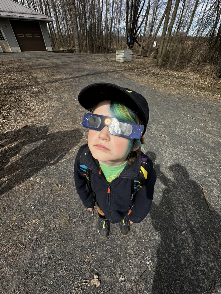 A wide angle photo of a kid wearing eclipse glasses. Her face in the centre of the frame and dirt visible below.  She's wearing a navy blue fleece jacket and a black baseball cap.