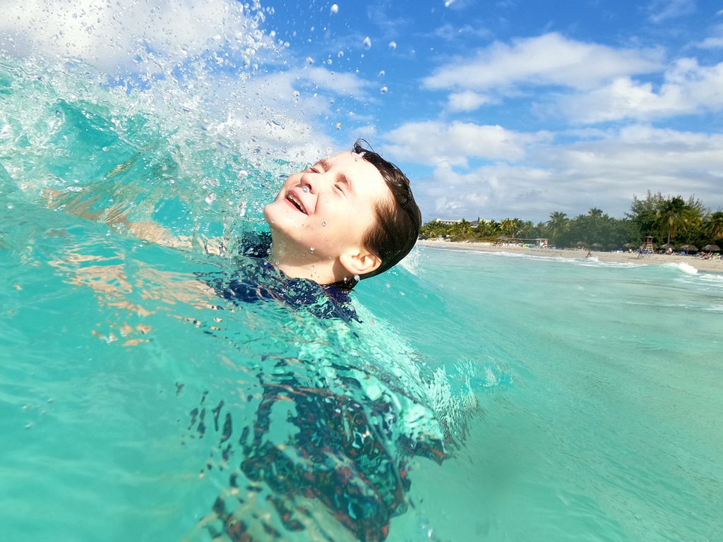 photo of a kid swimming in blue-green water caught in a wave. looking totally ecstatic. beach and green tree line visible in the distance