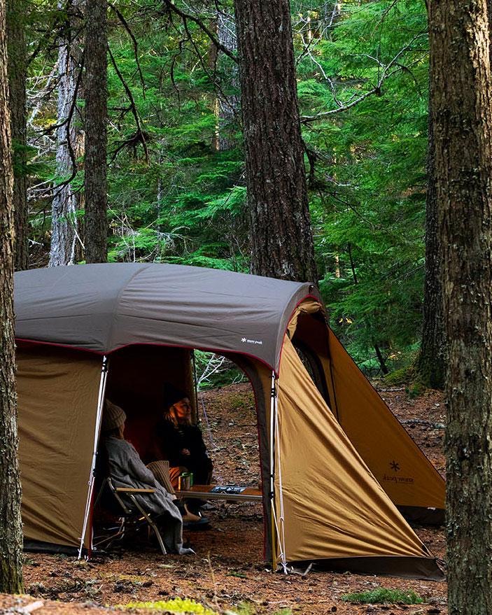 A photo of a tent in a forest. Pine trees visible and so are some people through an open door. The tent is light brown.