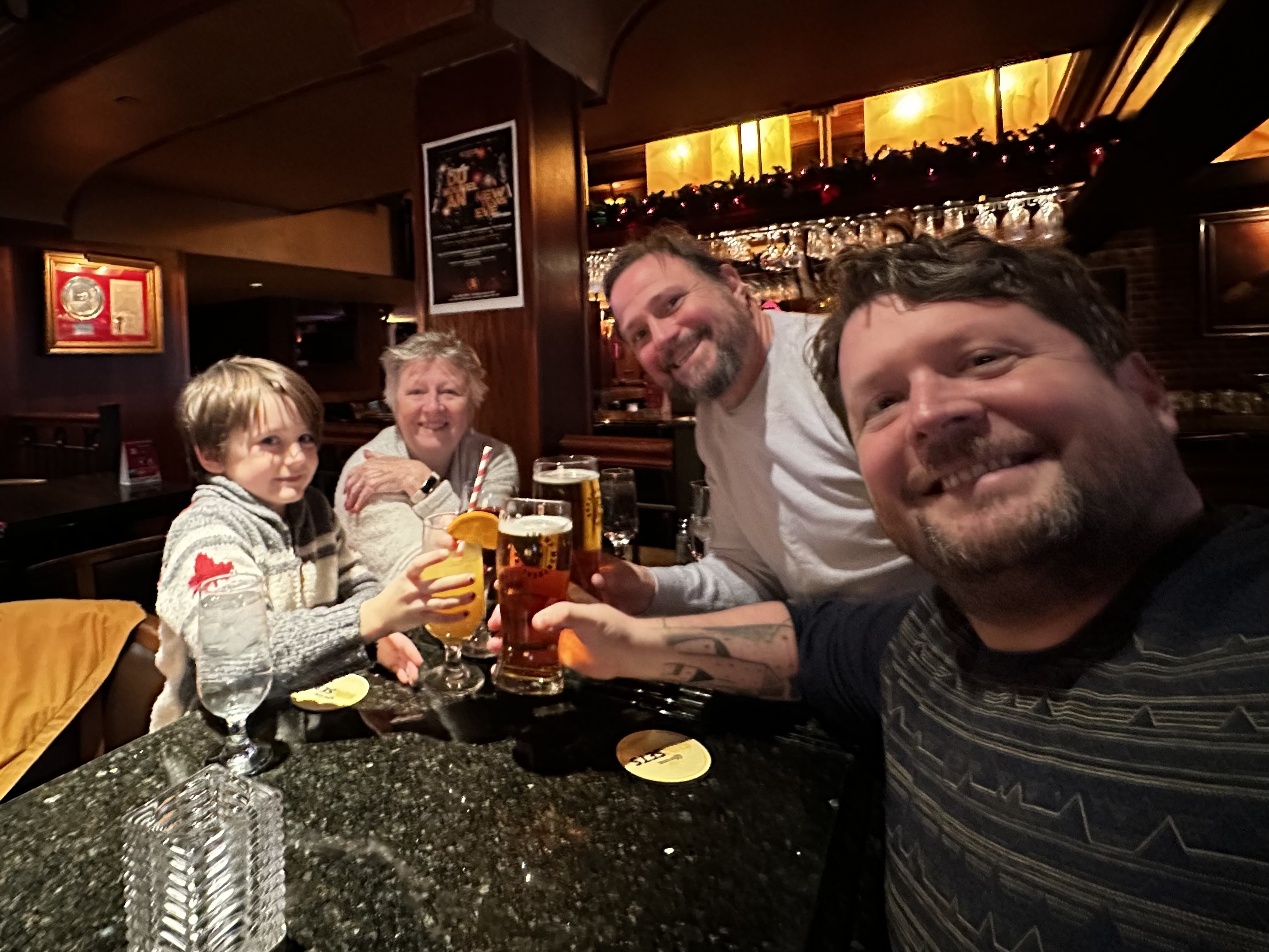 Wide angle selfie of four people at a restorant table. Dark wood room. Mid cheers with some drinks. 