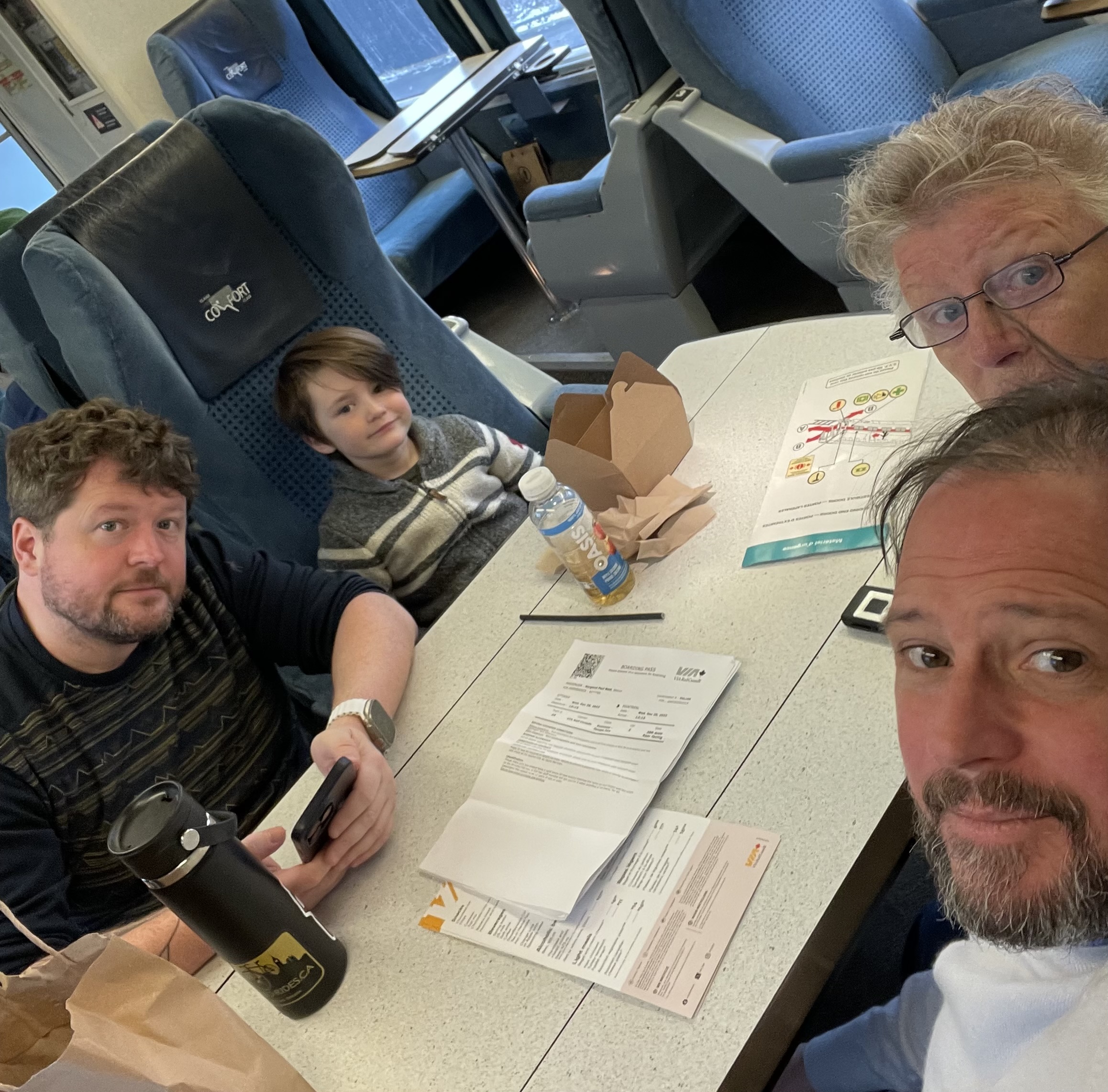 selfie of four people in a train. papers are visible on a table and they're sitting around it looking tired but excited for the vacation