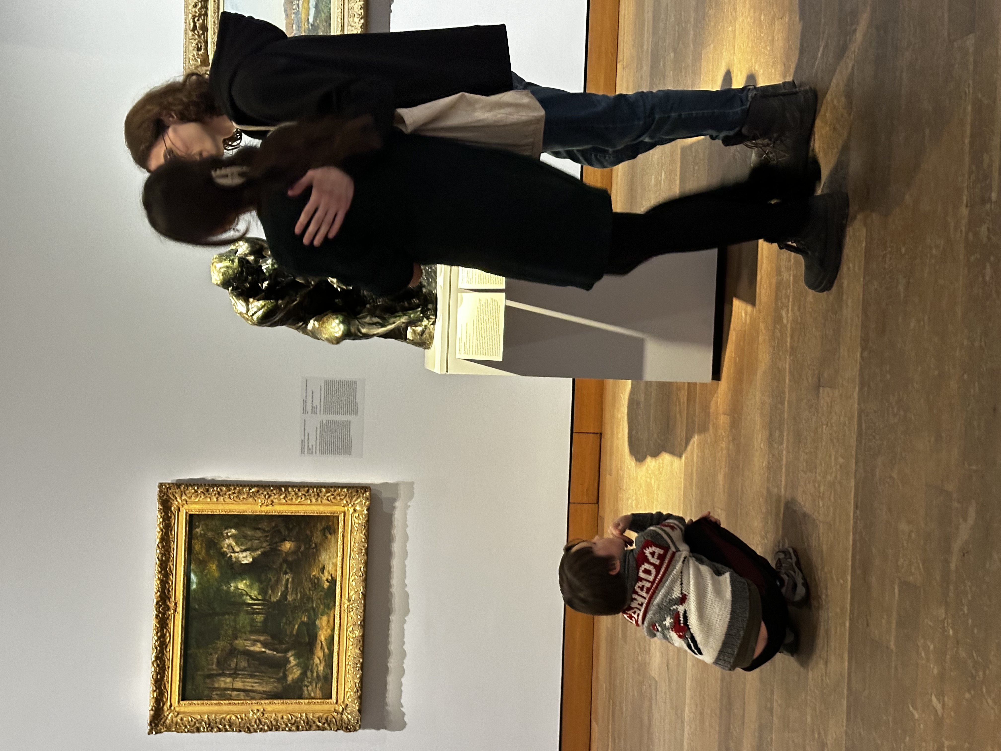 Photo taken in a museum hall. A smaller copy of Rodin’s The Thinker sits on a pedestal while a couple look on. A kid is crouched on the floor emulating the signature pose of the bronze statue. Fist under the chin, in thought.  