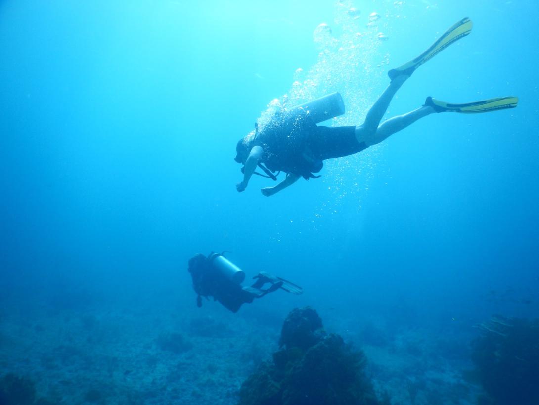Underwater photo of SCUBA divers. Two visible with some rocks. Bright blue water with divers in silhouette