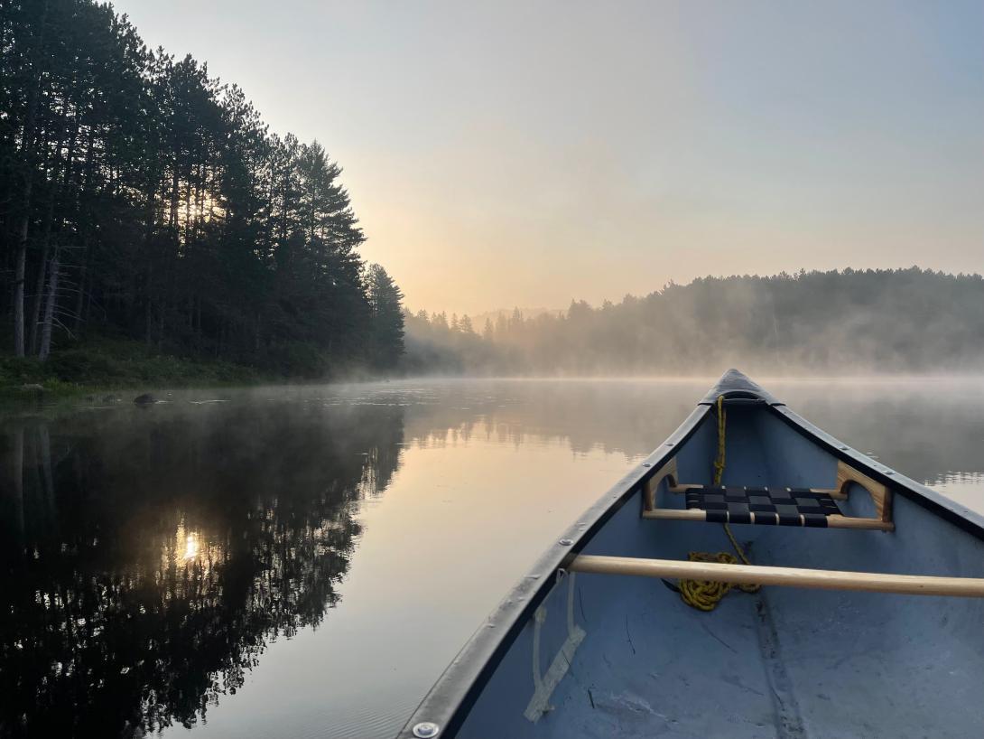 A still and foggy lake visible over the bow of a canoe. It's early morning and trees and a hill are visible over the lake.