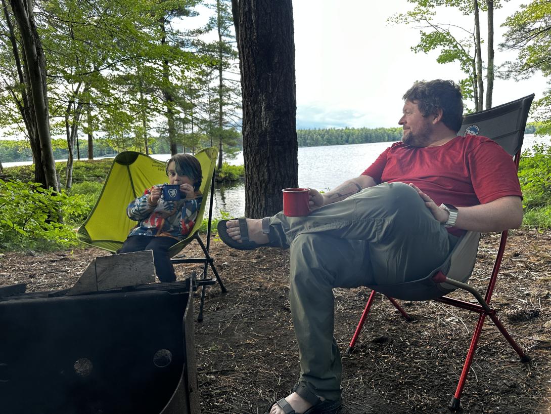 Camping at Frontenac 2024 Photo of a man sitting with legs crossed in a camping chair next to a kid in another chair. A lake and forest visible around them.